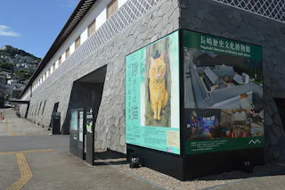 A stone building with a large sign featuring a painting of an orange cat and promotional images for the Nagasaki Museum of History and Culture, with a hillside and blue sky in the background.
