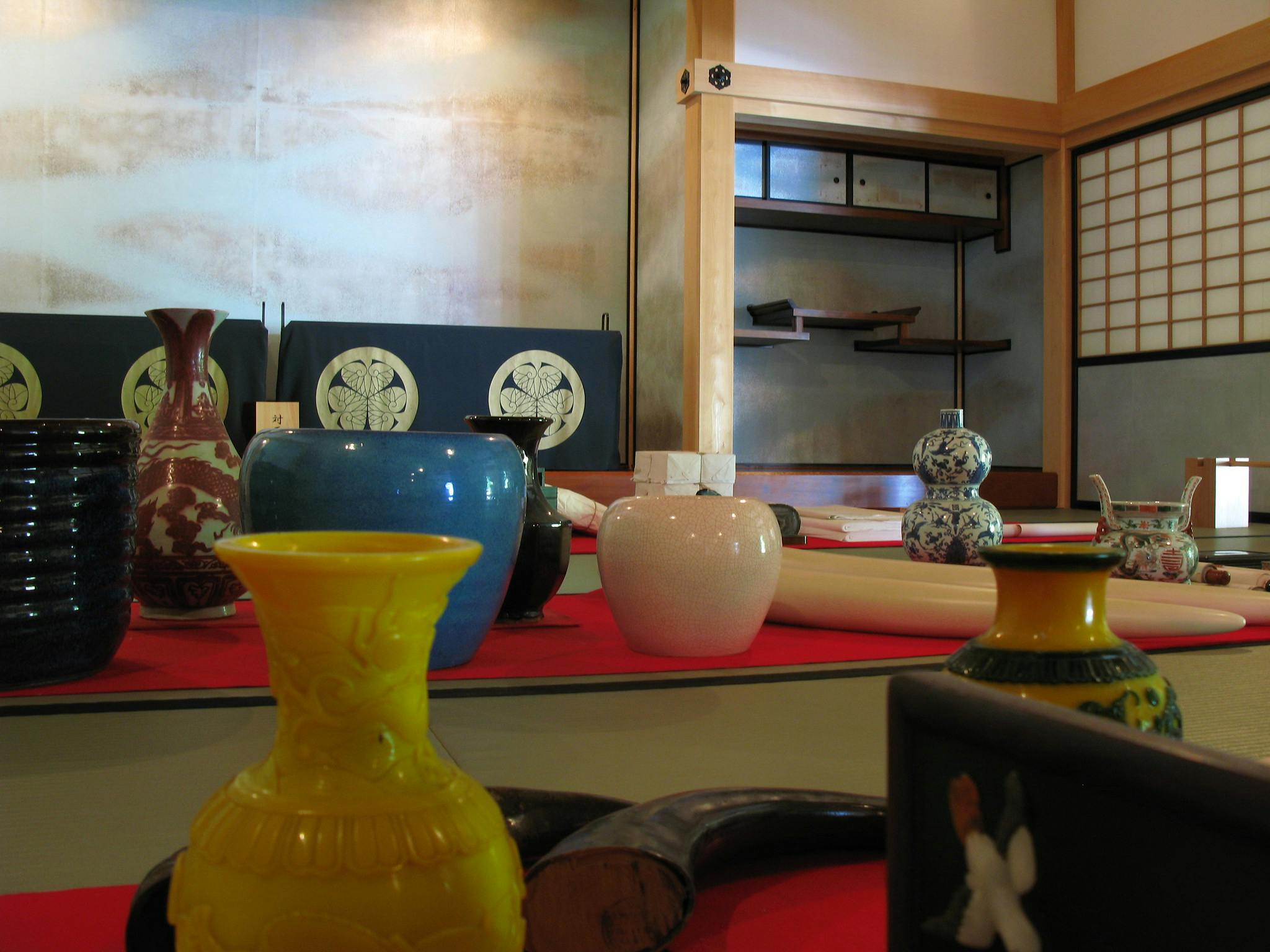 A variety of colorful ceramic vases and decorative objects are arranged on a red tablecloth in a traditional Japanese room with shoji screens and wooden shelves in the background.