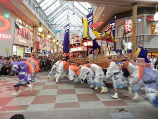 A group of people in traditional Japanese clothing dance energetically in front of a large decorated float inside a shopping arcade, watched by a crowd of onlookers. Colorful flags and lanterns adorn the float.
