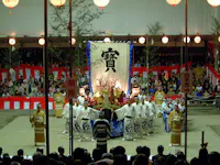 A large group of people in traditional Japanese clothing perform on an outdoor stage with red and white decorations, a large banner with kanji, and an audience watching the event at night.