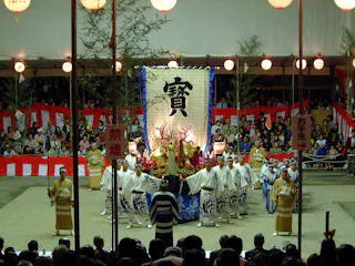 A large group of people in traditional Japanese clothing perform on an outdoor stage with red and white decorations, a large banner with kanji, and an audience watching the event at night.