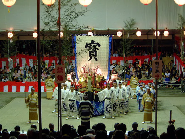 A large group of people in traditional Japanese clothing perform on an outdoor stage with red and white decorations, a large banner with kanji, and an audience watching the event at night.