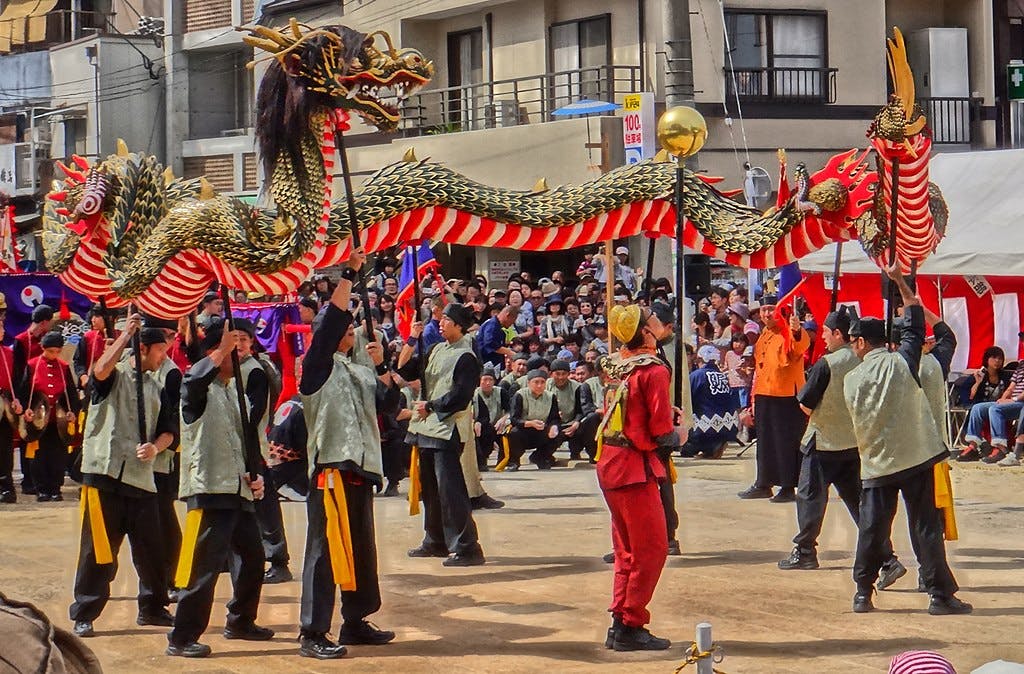 Performers carry a long, colorful dragon puppet on poles during a festival parade, with a crowd of people watching in the background. The event takes place outdoors in an urban setting.