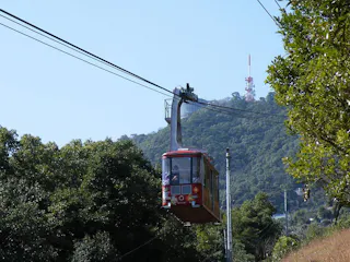 A red cable car travels above green trees on a hillside, with a large hill and a tall radio or TV tower in the background under a clear blue sky.