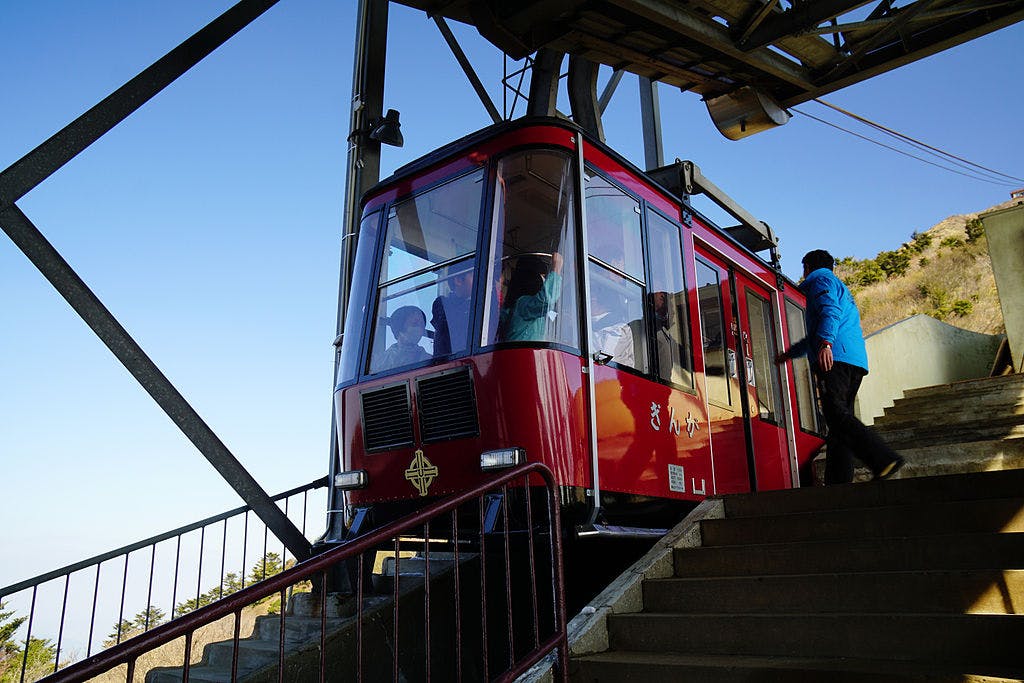 A red cable car waits at a station platform as a person in a blue jacket prepares to board, with metal railings and stairs in the foreground and a clear blue sky above.