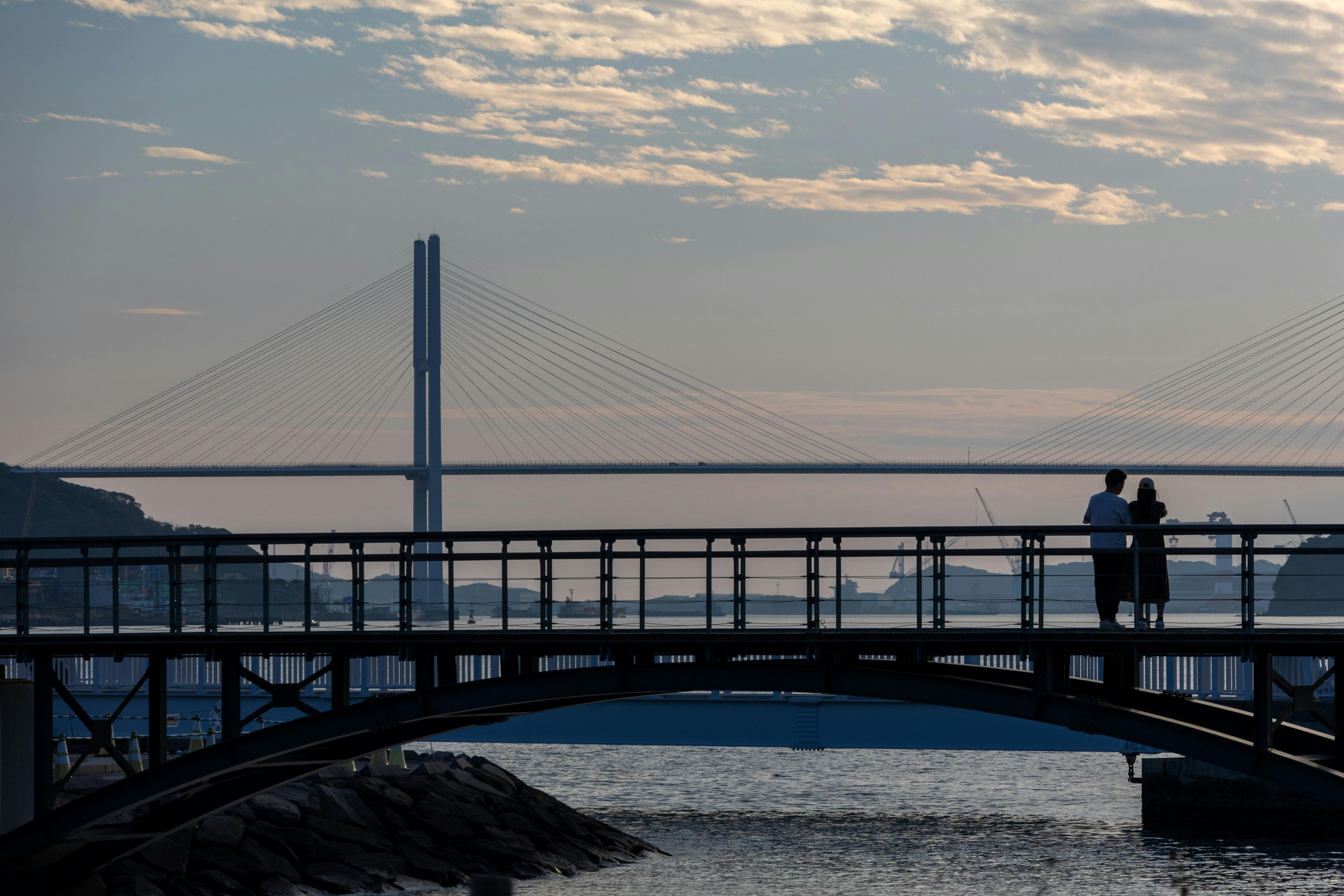 Two people stand on a pedestrian bridge over water at sunset, with another large cable-stayed bridge and a partly cloudy sky visible in the background.