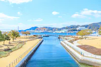 A clear blue canal flows under a small stone bridge, flanked by manicured lawns and walkways. In the background are mountains, cranes, and buildings near the waterfront under a sunny sky with scattered clouds.