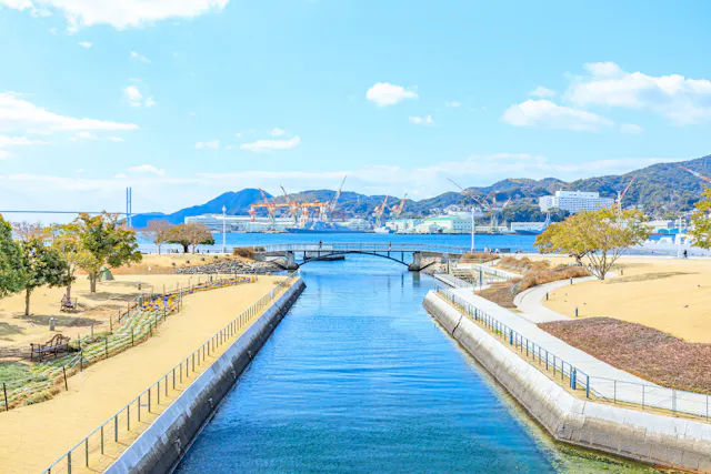 A clear blue canal flows under a small stone bridge, flanked by manicured lawns and walkways. In the background are mountains, cranes, and buildings near the waterfront under a sunny sky with scattered clouds.