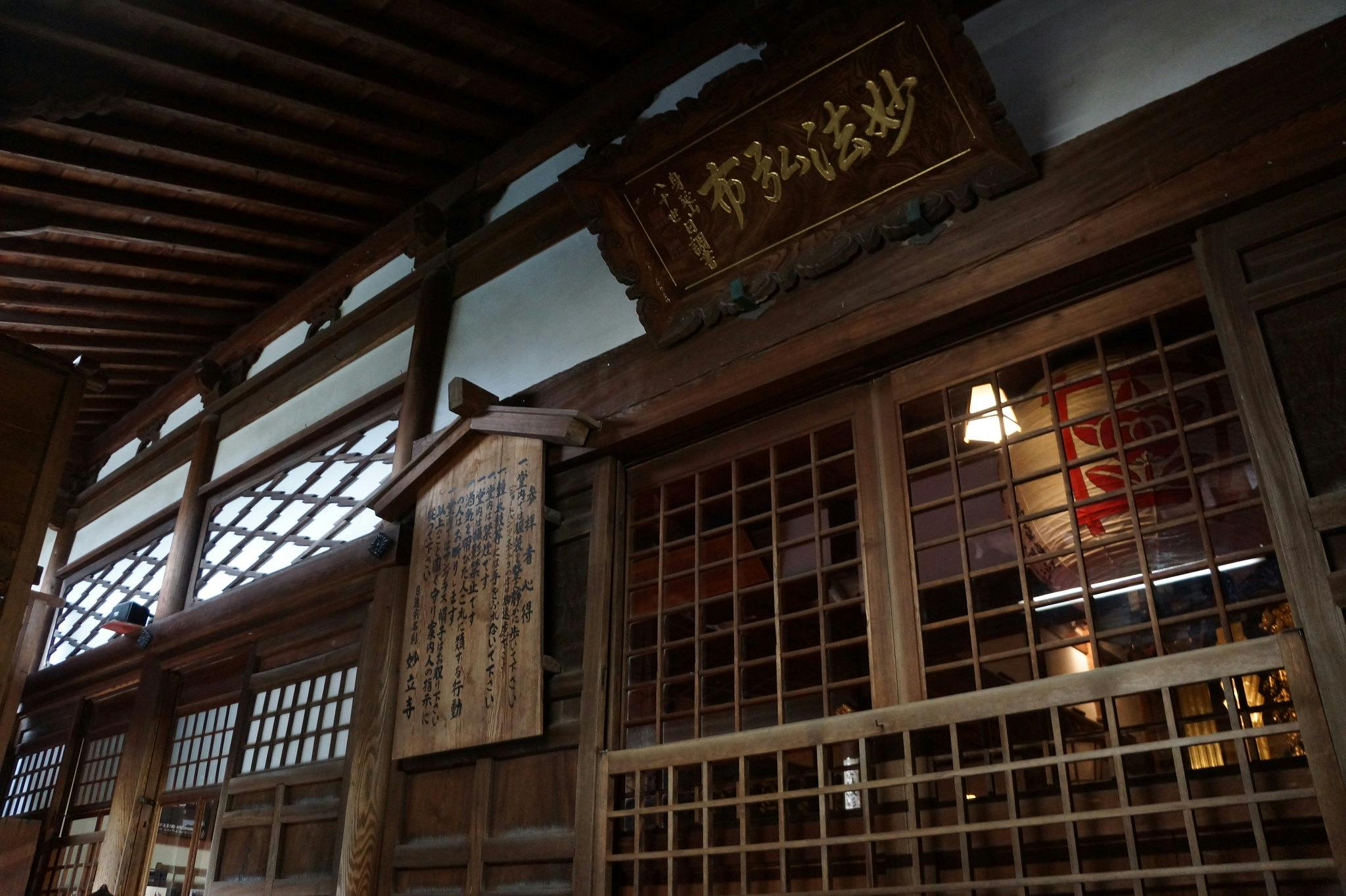 Wooden interior of a traditional Japanese building with sliding lattice doors, wooden plaques displaying Japanese calligraphy, and soft indoor lighting.