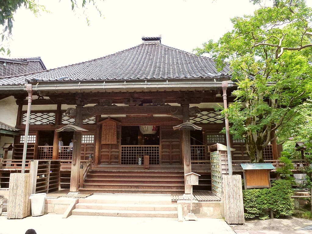 A traditional Japanese wooden temple with a tiled roof, open entrance, and steps leading up to the doorway, surrounded by green trees and a peaceful outdoor setting.