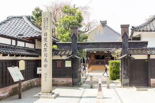 Traditional Japanese temple entrance with a tall stone pillar inscribed with Japanese characters, wooden gate, tiled roofs, and a path leading to the temple building, surrounded by trees and informational signs.