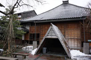 A traditional Japanese wooden building with a sloped, tiled roof. Snow is gathered at the base and on the ground. A wooden triangular structure shelters the entrance, and trees are visible nearby.