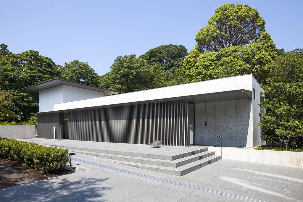 A modern, minimalist building with concrete walls and horizontal slatted panels, set among lush green trees, featuring wide steps leading to the entrance under a clear blue sky.