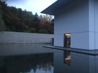A modern white building with clean lines sits surrounded by calm reflective water, bordered by a stone wall and autumn trees in the background under a cloudy sky.