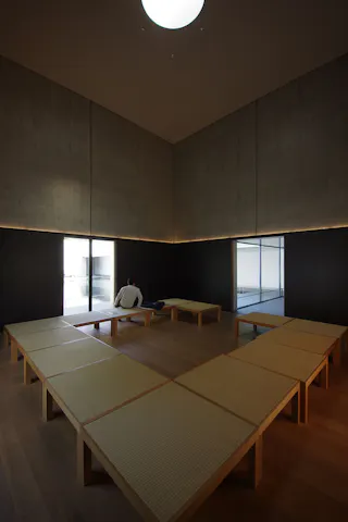 A person sits alone on a wooden bench in a spacious, minimalist room with high ceilings, concrete walls, and large windows that let in natural light. The benches form a U-shape around the room.