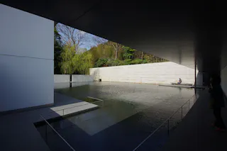 A modern outdoor courtyard with a reflective pool, white walls, and minimalist architecture. Trees are visible in the background, and a person sits by the water's edge in the sunlight.