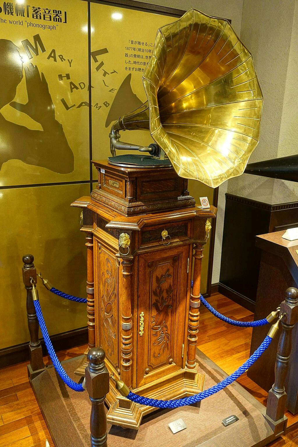 A vintage gramophone with a large brass horn sits atop an ornate wooden cabinet, displayed behind blue ropes in a museum setting with wood floors and informational panels in the background.