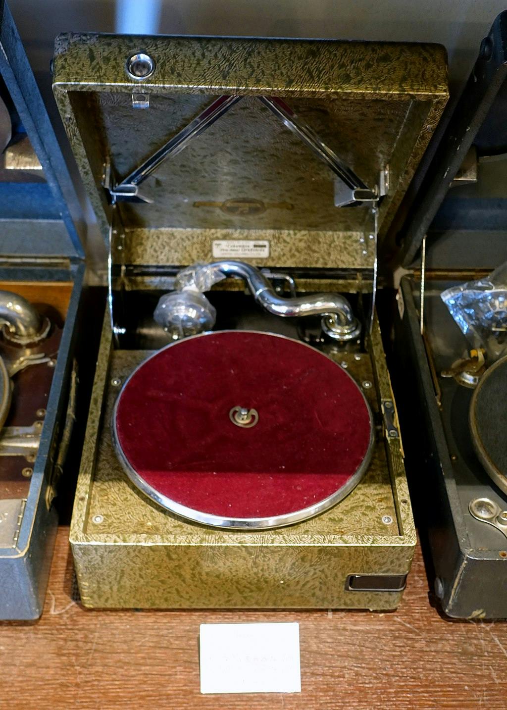 An antique portable gramophone with a hinged lid open, revealing a red felt turntable, a metal tonearm, and needle, displayed on a wooden surface.