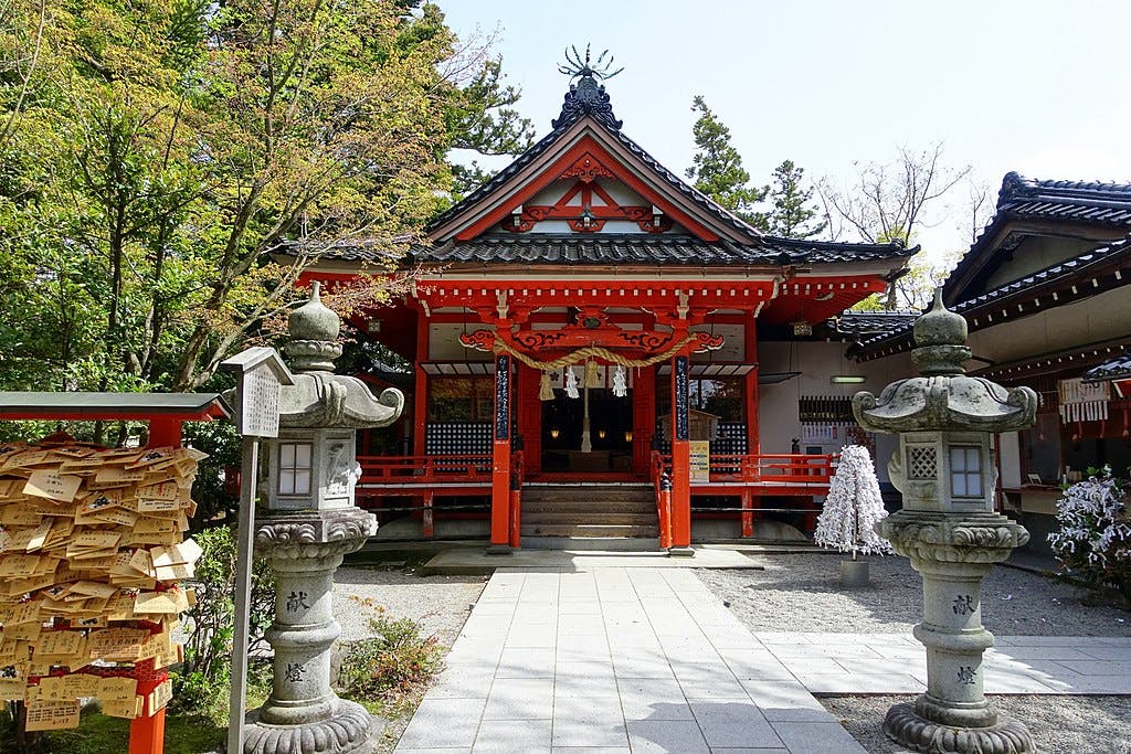 A traditional Japanese shrine with a red and black roof, stone lanterns, and wooden prayer plaques displayed on a stand. Trees and other smaller buildings surround the shrine.