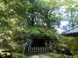 A small cave entrance covered in moss and greenery sits beneath dense trees, with a bamboo fence in front and a traditional tiled roof visible to the right. Sunlight filters through the foliage.
