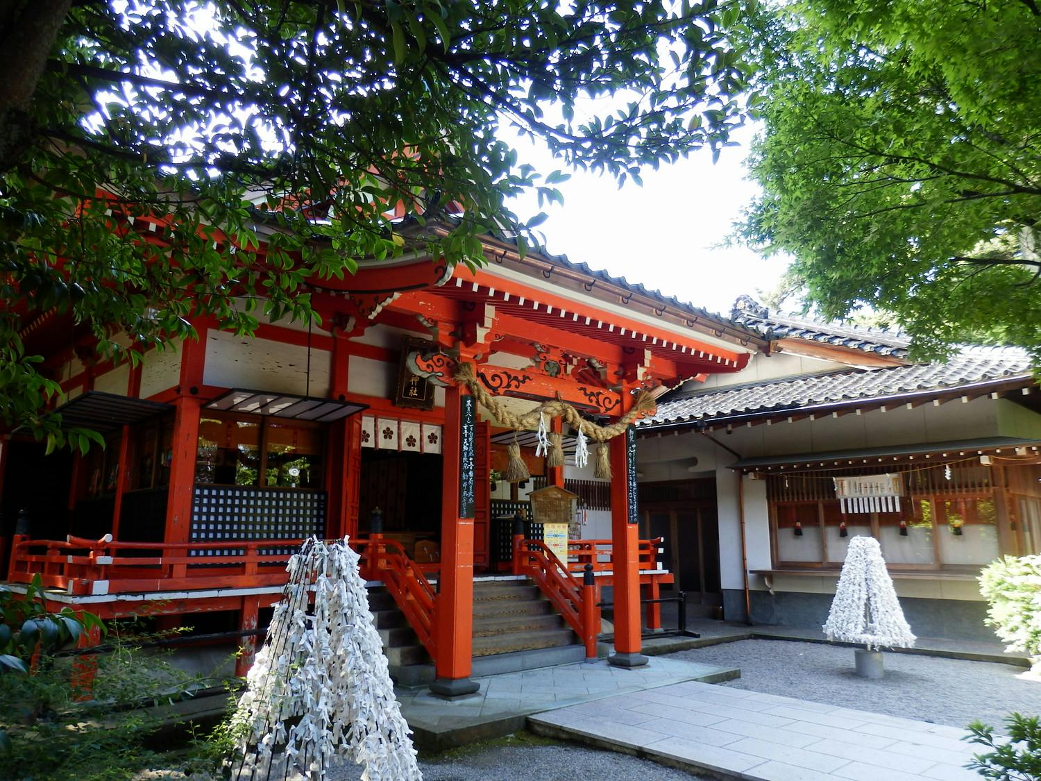 A traditional Japanese Shinto shrine with red and white wooden architecture, surrounded by lush green trees. There are steps leading up to the entrance and decorative paper fortunes tied onto stands in the courtyard.