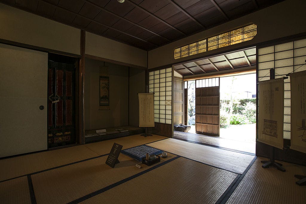 A traditional Japanese tatami room with sliding shoji doors open to a sunlit garden, tea utensils on the floor, hanging scrolls, and soft natural light filling the space.