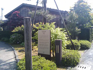 A traditional wooden Japanese house surrounded by lush greenery, with a tall wooden signpost and an information board written in Japanese near the entrance. Sunlight brightens the scene.