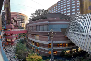 A wide view of a modern multi-story shopping complex with colorful buildings, large windows, a garden courtyard below, and a covered walkway connecting two sections of the complex.