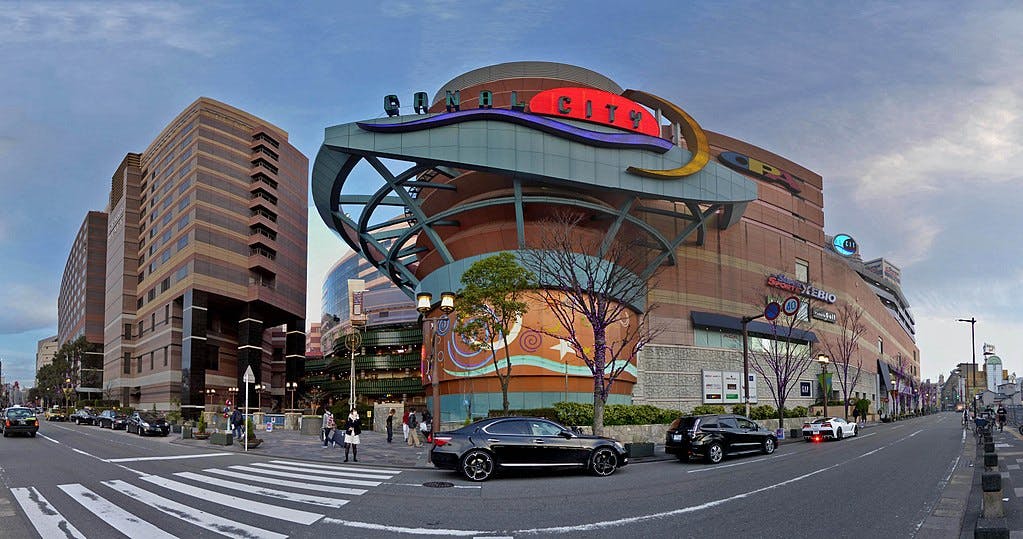 A city street scene with a large modern shopping mall called Canal City, featuring colorful, curving architecture, trees, cars, and tall buildings under a partly cloudy sky.