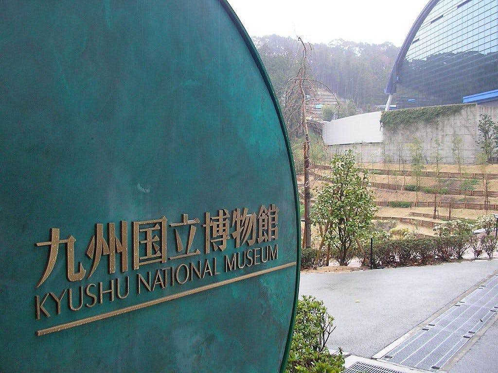 A large round sign reading "Kyushu National Museum" in Japanese and English stands near a modern museum building with a curved glass roof, surrounded by trees and a pathway.