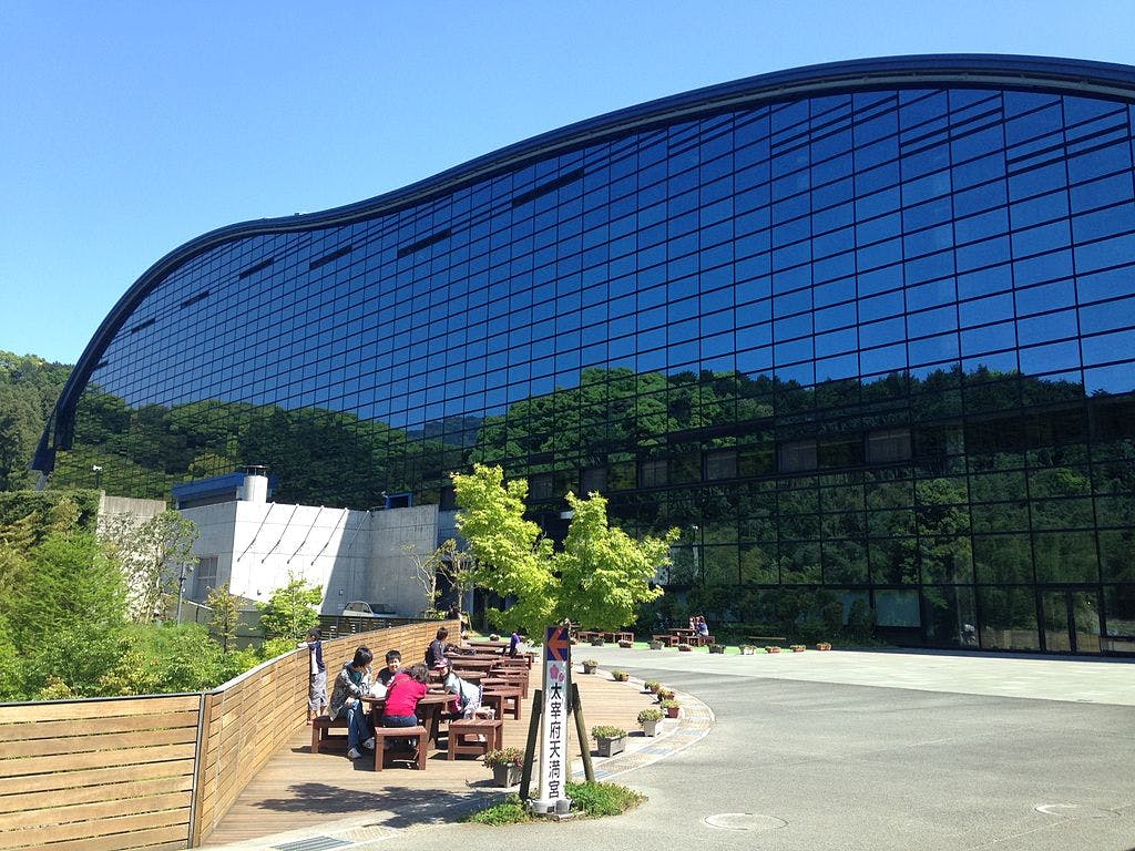 A group of people sits at picnic tables outside a large, modern, curved glass building reflecting trees and blue sky, with green foliage and a clear, sunny day.