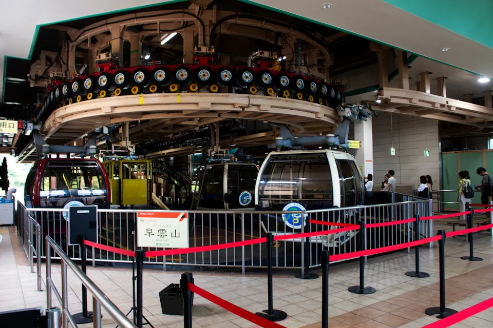 A cable car station with colorful gondolas, including red, yellow, and white ones, parked and waiting. Red ropes guide the queue area, and a few people are visible in the background. An overhead mechanism for the cable cars is visible above the gondolas.