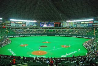 A wide view of an indoor baseball stadium with a green artificial field, players positioned for a game, scattered spectators in the stands, and advertisements on the walls under a large domed roof.