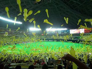 A large crowd in a stadium releases yellow balloons into the air, filling the field and stands with flying balloons under bright stadium lights and a domed roof.