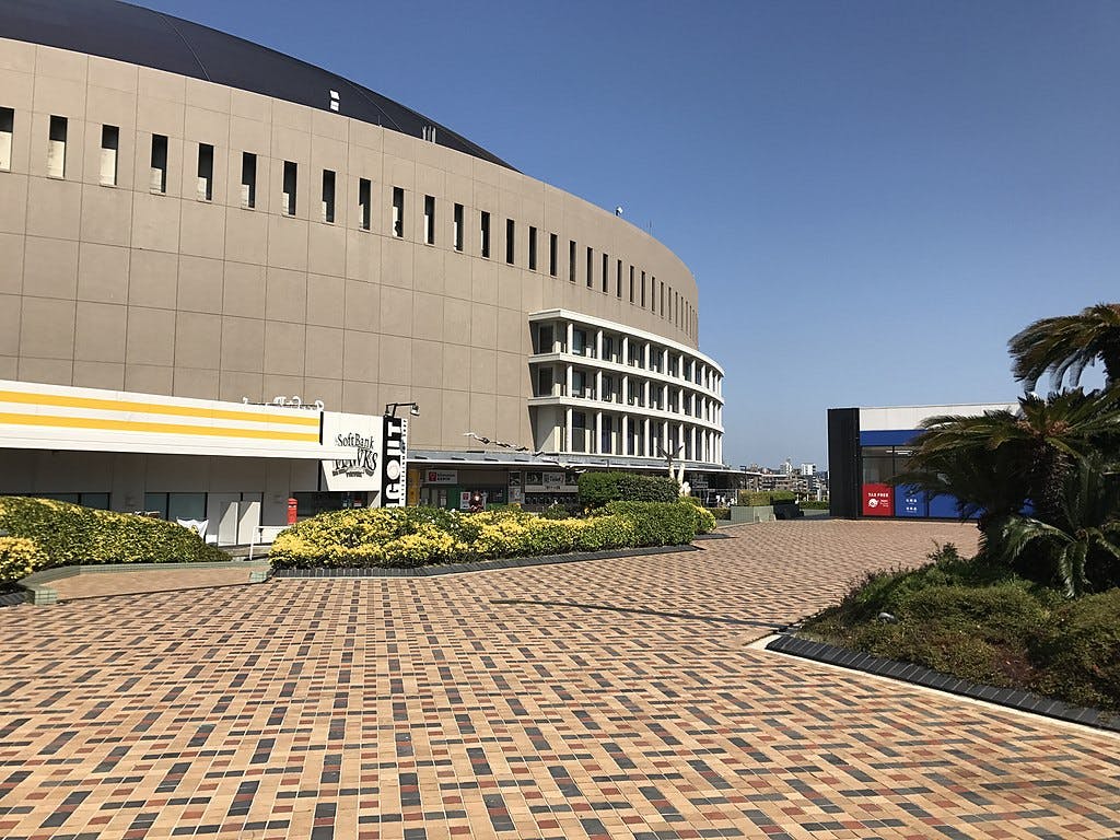 A large circular building with beige walls stands next to a patterned brick plaza, bordered by green bushes and palm trees under a clear blue sky. Some small shops and signs are visible near the building.