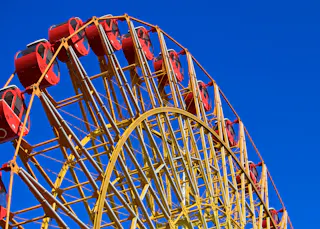 A close-up view of a yellow Ferris wheel with red gondolas set against a clear, bright blue sky. The intricate metal framework of the Ferris wheel is prominently visible.