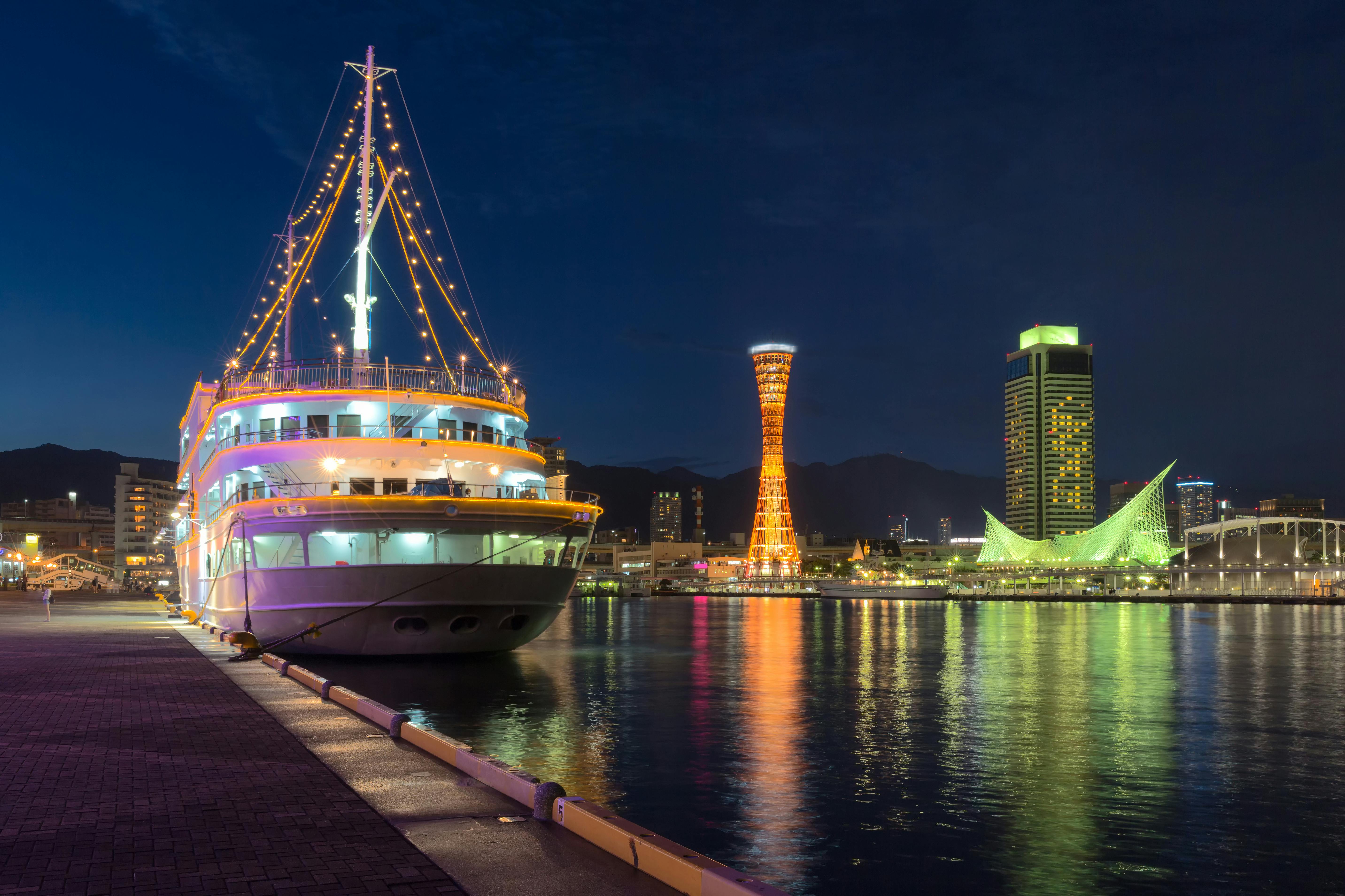 A brightly lit cruise ship docked at a harbor at night, with city buildings and illuminated towers reflecting on the calm water in the background.