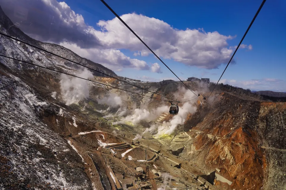 A scenic view of a mountain landscape with steam rising from the ground, showcasing a volcanic area. Two cable cars are suspended on cables running across the scene. Snow patches cover parts of the terrain, and the sky is partly cloudy with a blue backdrop.