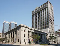 A large, historic stone building with columns stands in front of two modern high-rise buildings under a clear blue sky. People are walking on the sidewalk and trees are visible along the street.