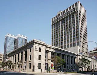 A large, historic stone building with columns stands in front of two modern high-rise buildings under a clear blue sky. People are walking on the sidewalk and trees are visible along the street.