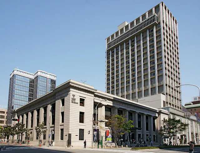 A large, historic stone building with columns stands in front of two modern high-rise buildings under a clear blue sky. People are walking on the sidewalk and trees are visible along the street.