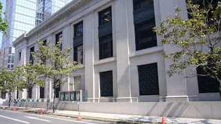 A large, white stone building with tall windows and columns stands along a city street lined with young trees and orange traffic cones. Modern glass buildings are reflected in the background.