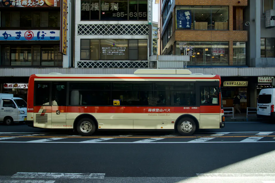 A red and white bus is parked on a city street. Behind the bus are buildings with various signs and storefronts. The street appears calm, and a few other vehicles, including a white van, are visible next to the bus.