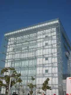 A modern building with a glass exterior and visible structural framework, with trees and a clear blue sky in the background.