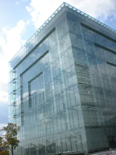 A modern glass building with a transparent, geometric facade reflects clouds and the sky. A small tree stands in front of the structure, emphasizing the building's sleek, angular lines and minimalist design.