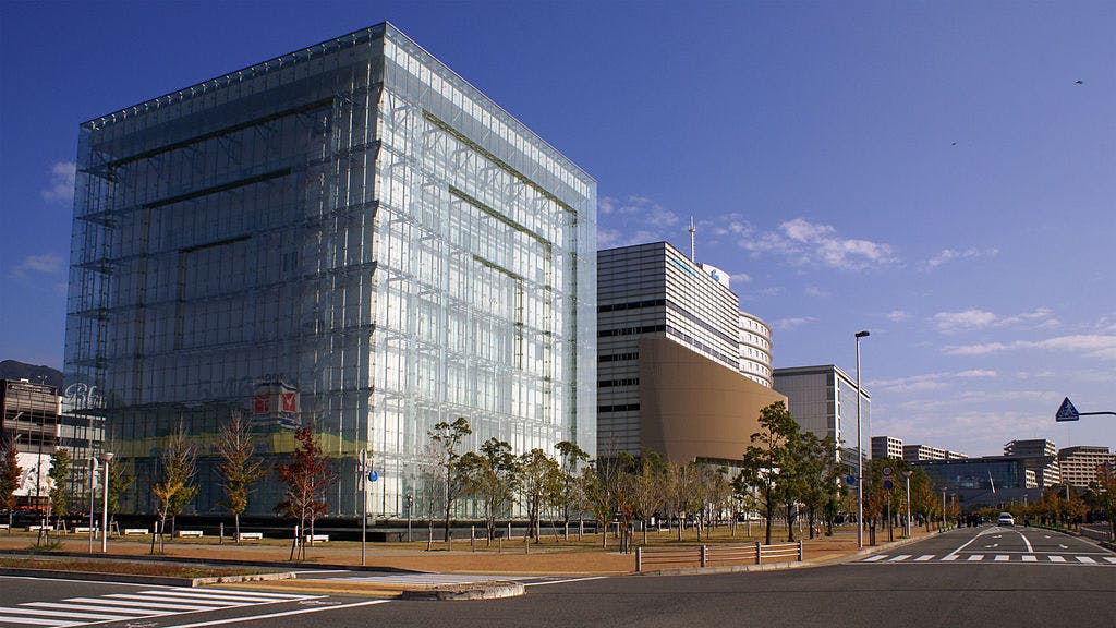 A modern glass office building with trees and a wide road in front, set under a clear blue sky with some scattered clouds. Other buildings are visible in the background.