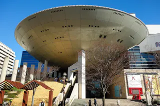 A large, circular, UFO-shaped structure covers the entrance of a modern building with glass and stone walls. People walk up wide outdoor steps under a clear blue sky. Leafless trees and signs are visible.