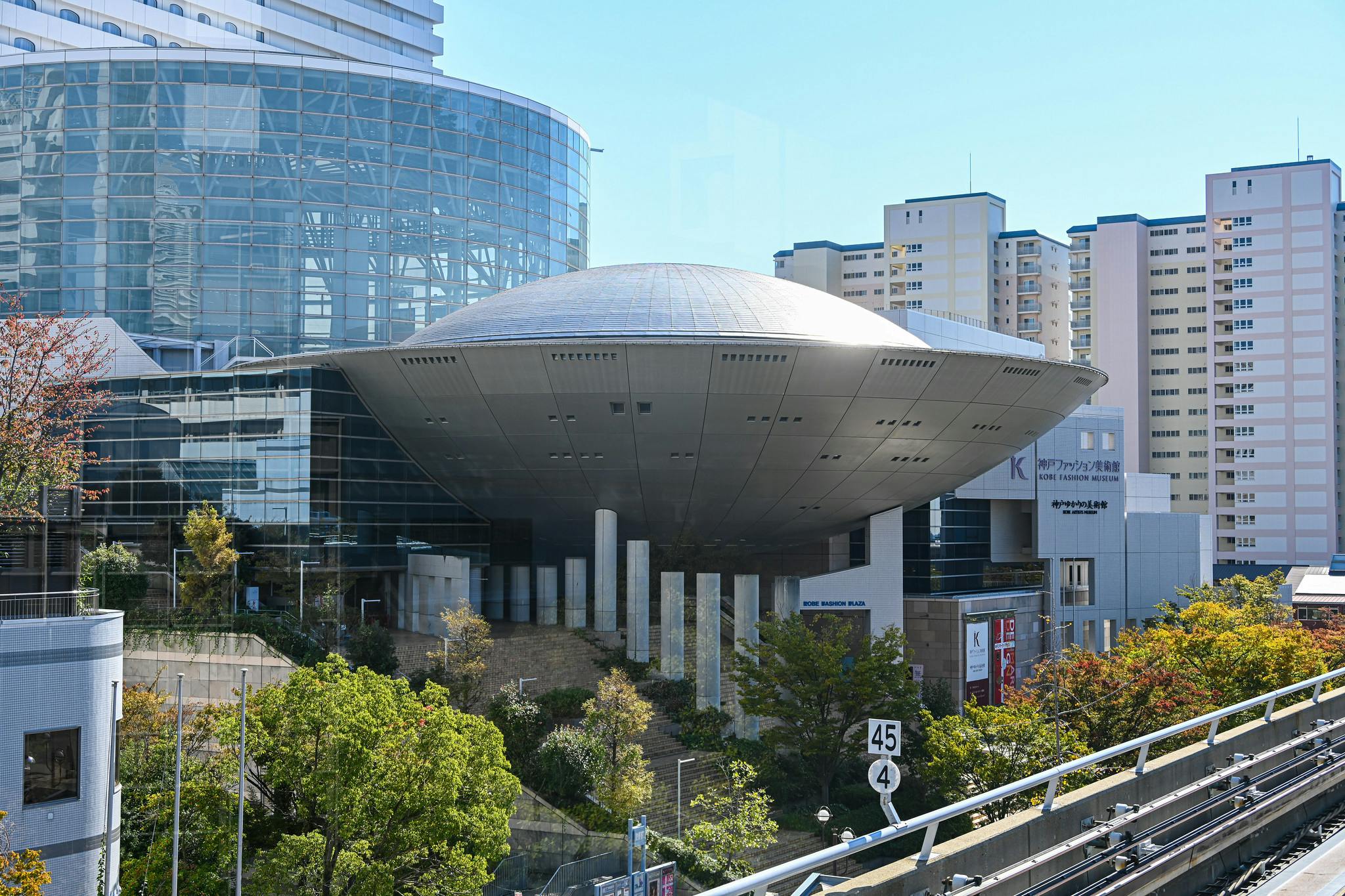 A modern building with a large, silver, UFO-shaped structure elevated on columns, surrounded by trees, other buildings, and high-rise apartments in an urban area.