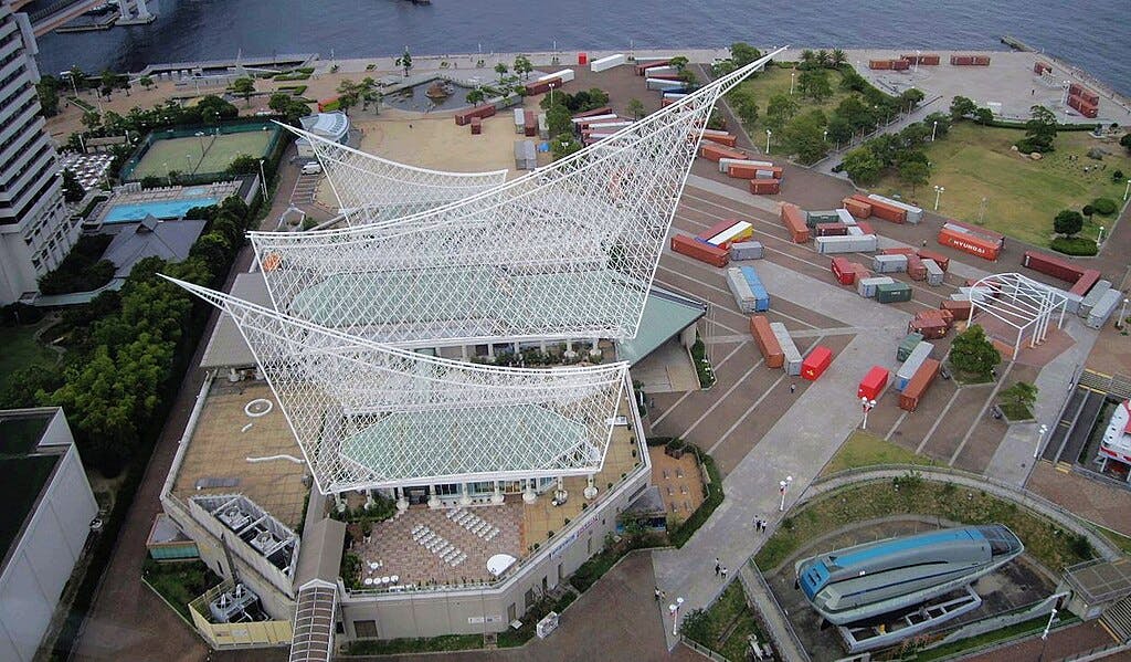 Aerial view of a modern waterfront building with angular, white steel structures resembling sails, surrounded by trees, walkways, and colorful shipping containers near a body of water.