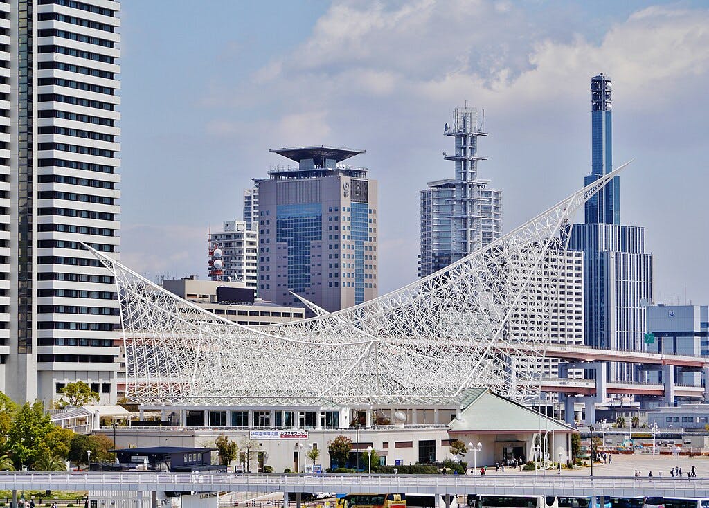 A modern cityscape featuring tall skyscrapers and the Kobe Maritime Museum with its distinctive white, sail-shaped steel structure in the foreground under a partly cloudy sky.
